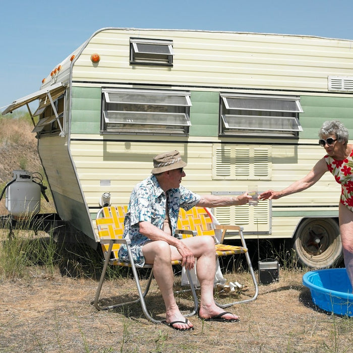 A photo of an old couple camping outdoors next to a caravan with a lady in a shallow pool.