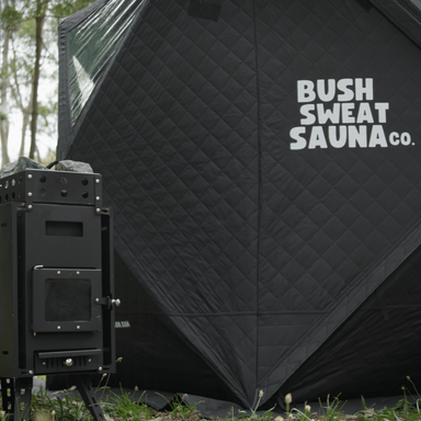 A photo of a 4-6 person portable sauna tent sitting next to a stove with rocks on top of it.