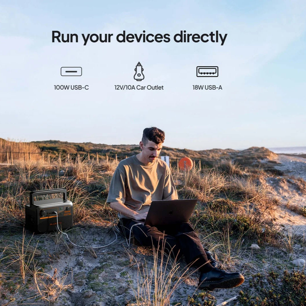 A photo of a man sitting on grass, charging his laptop which is connected to a Bluetti B80P power station expansion battery.