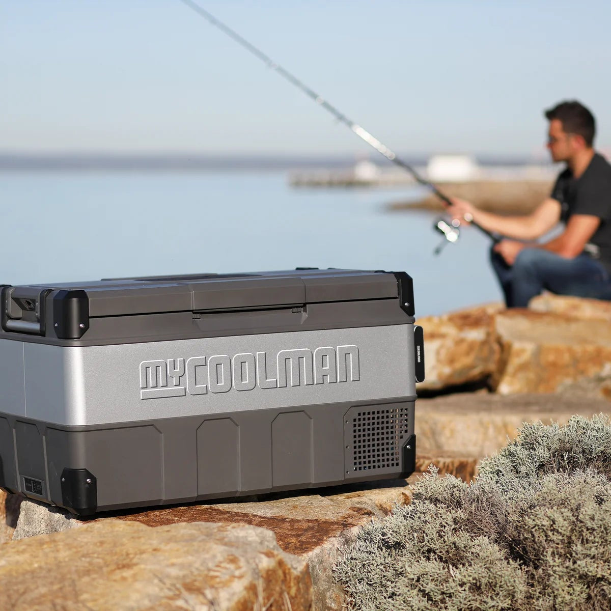 A photo of a MyCOOLMAN 105L Portable Car and Camping Fridge outdoors with a man fishing in the background.