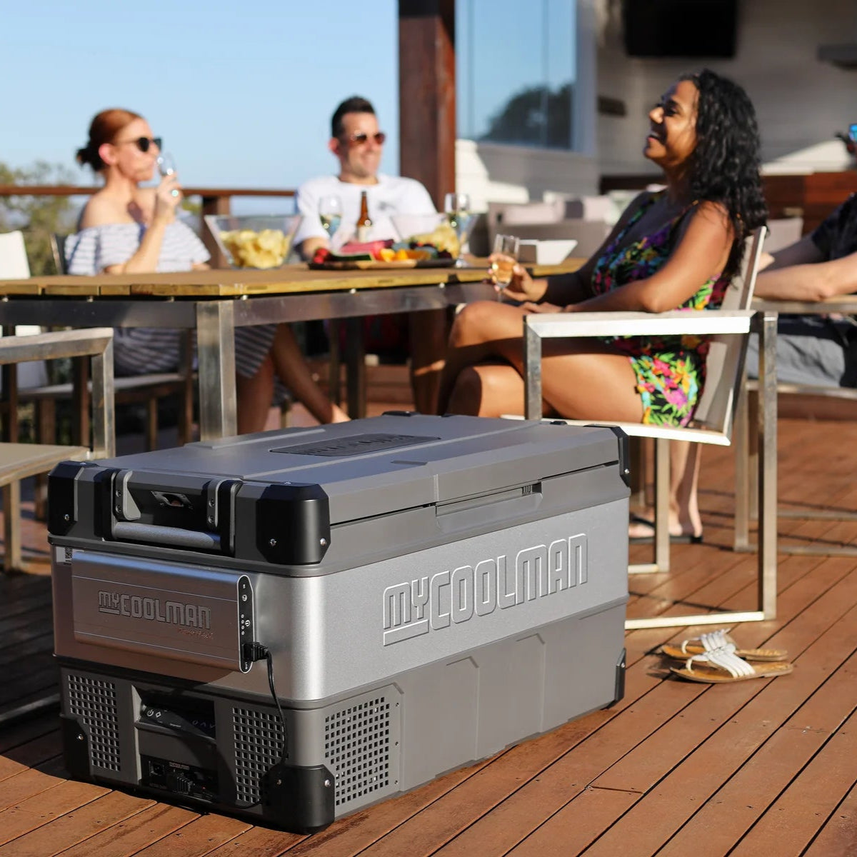 A photo of a MyCOOLMAN 105L Portable Car and Camping Fridge on a deck with a group of people enjoying beverages.