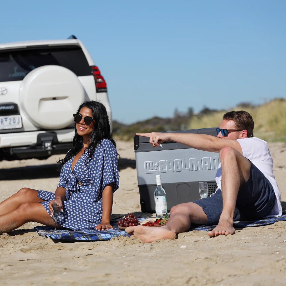 A photo of a man and a woman next to a MyCOOLMAN 69L Dual Zone Portable Car and Camping Fridge enjoying a drink and food.