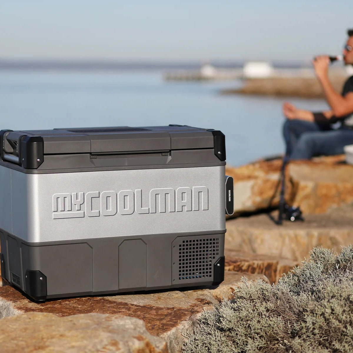 A photo of a MyCOOLMAN 73L Portable Car and Camping Fridge outdoors with a man drinking a beverage in the background.