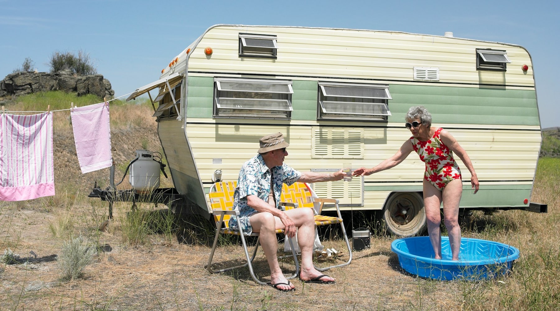 A photo of an old couple camping outdoors next to a caravan with a lady in a shallow pool.