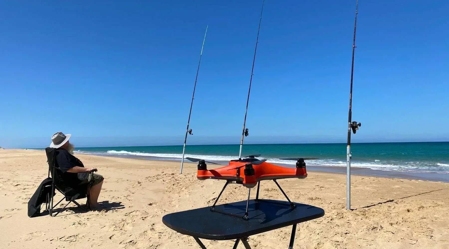 A man sitting on a beach with fishing rods and a swellpro fishing drone.