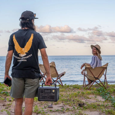 A picture of a man holding a bluetti eb3a 600w portable power station next to water, with a woman looking over on a deck chair.
