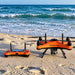 SwellPro Fisherman FD3S orange fishing drone and remote controller sitting on a sandy beach with ocean waves in the background.