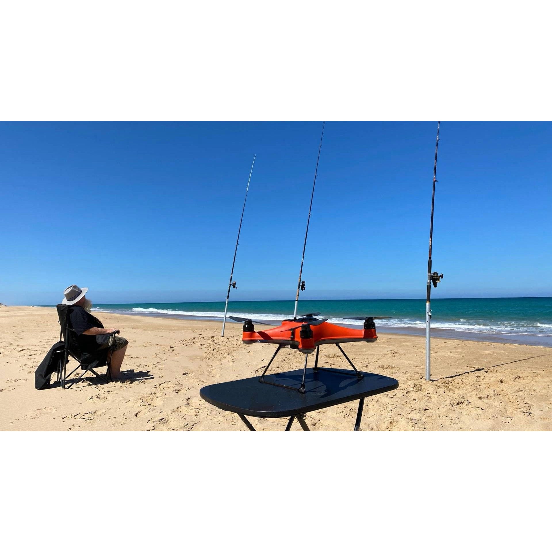 SwellPro Fisherman FD1S fishing drone set up on a table on an Australian beach beside three surf rods, with an angler seated in a chair watching the water.