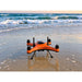 SwellPro Fisherman FD3S fishing drone standing on wet sand at the water's edge at sunset, with surf breaking behind it.